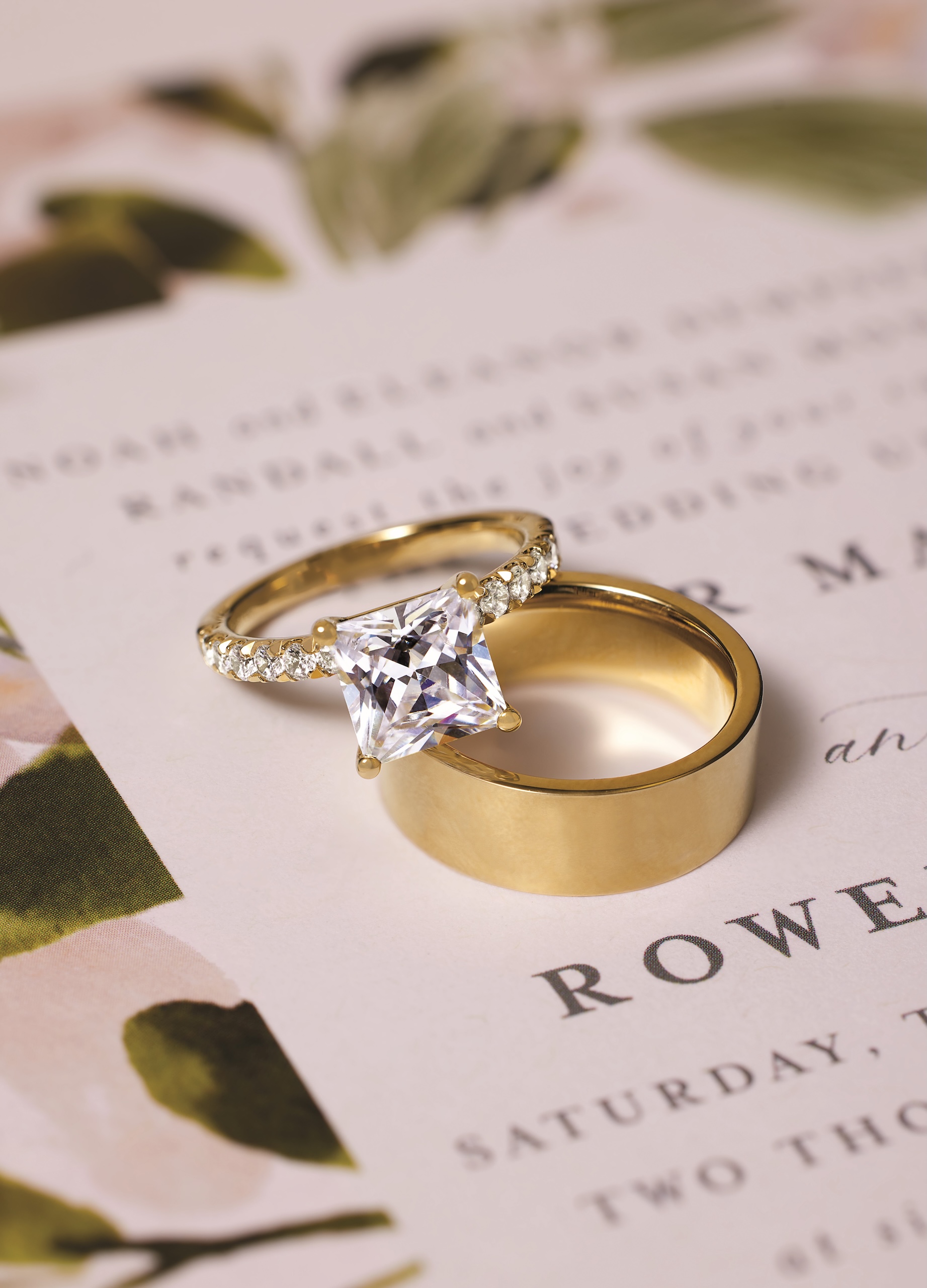 A beautiful bridal set, featuring a princess cut diamond engagement ring and a classic gold wedding band, resting on a wedding invitation, symbolizing the start of a new journey with Catawba Island Jewelers.